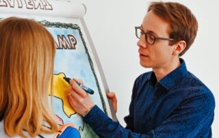 Tom Rescke und unbekannte Frau zeichnen gemeinsam eine Landkarte auf einem Flipchart.
