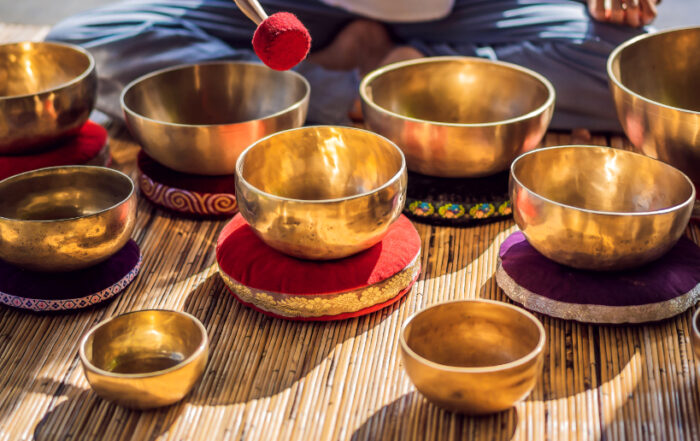 Nahaufnahme mehrerer goldener, tibetischer Klangschalen auf Kissen, die auf einer Bambusmatte für eine Klangtherapie oder Meditation arrangiert sind. Eine Person hält einen Schlägel bereit.